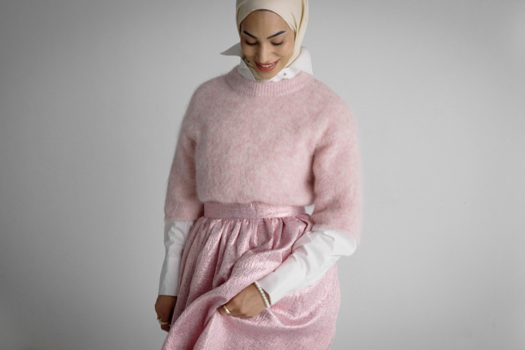 Model looking down gently at her clothes, wearing a pink long skirt, a pink woolen oversized sweater and matching hijab.